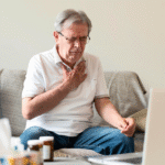 Elderly man sitting on a couch, clutching his chest with a pained expression, possibly experiencing chest pain or difficulty breathing, near medication bottles.