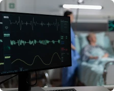 Heart rate monitor displaying vital signs beside a hospital bed with a patient and healthcare worker in the background