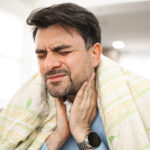 A man wrapped in a blanket, holding his throat, showing signs of discomfort from ear pain and sore throat.