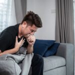 A man sitting on a couch, mouth open and coughing, showing symptoms of an Upper Respiratory Infection.