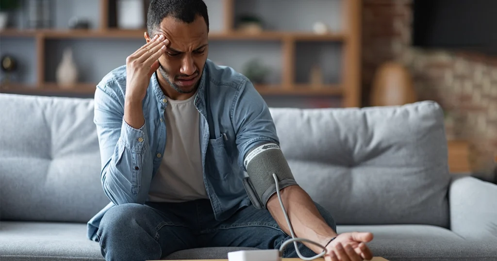Man sitting on a sofa measuring blood pressure at home, looking stressed and dizzy.